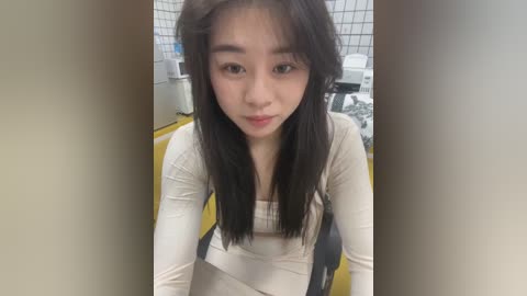 Media: Video of a young East Asian woman with long black hair, wearing a beige sweater, in a clean, white-tiled bathroom with a yellow floor. She appears serious and slightly pouty.