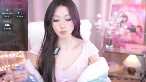 Media: Video of a young Asian woman with long black hair, wearing a light pink shirt, sitting on a white chair, indoors with a soft, pink-lit background.