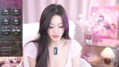 Media: Video of an East Asian woman with long, dark hair, wearing a pink top and necklace, in a soft-focus bedroom with pastel decor.