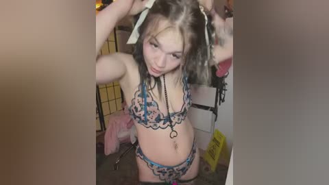 Media: Video of a young woman with light skin, long dark hair, wearing a blue and white floral bikini, standing in a dimly lit room with a bed and yellow caution tape in the background.