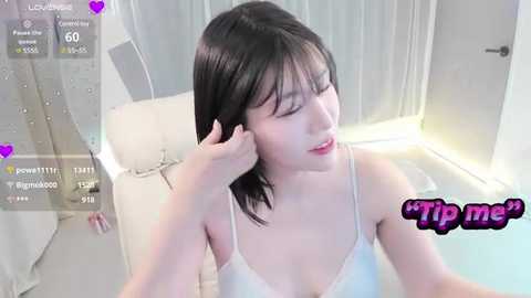 Media: Video of an Asian woman with straight black hair, wearing a white bra, sitting in a bathtub. She has a fair complexion and a soft, serene expression. The background shows a modern bathroom with white tiles and a glass shower door.