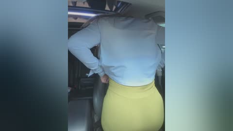 Media: Video of a woman standing in a car, wearing a white blouse and tight yellow skirt. Her back is to the camera.