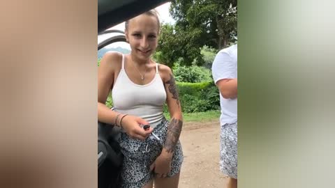 Media: Video of a smiling woman with light skin and brown hair in a white tank top and patterned shorts, standing next to a car in a lush green environment.