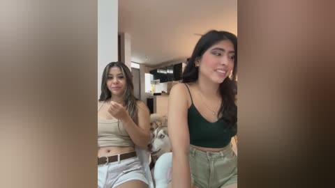 Media: Video of two young women in casual attire: one in a beige crop top and white shorts, the other in a green crop top and high-waisted shorts. Background features a modern kitchen with beige walls and dark cabinets.