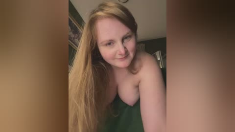 Media: Video of a smiling, topless, fair-skinned woman with long, straight, blonde hair, leaning forward with medium-sized breasts, in a dimly lit room with a framed painting on the wall.