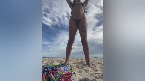 Media: Video of a bikini-clad woman standing on sandy beach, with colorful beach towel near her feet, under a partly cloudy sky.