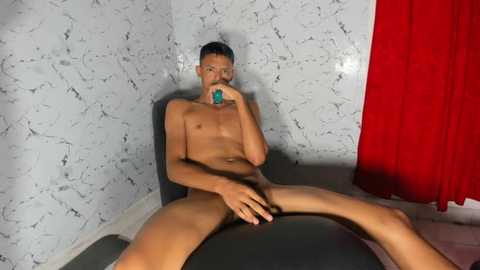 Media: Video of a naked, muscular Asian man with short black hair, holding a blue vibrator, sitting on a black chair in a room with white marble-patterned walls and red curtains.