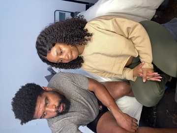 Media: Video of a Black woman with curly hair and a beige sweater, sitting on a bed with a Black man in a grey shirt, both looking relaxed and happy.