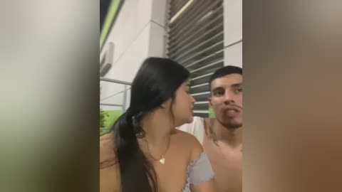 Media: Video of a young woman with long black hair, tan skin, and a light blue off-shoulder top, kissing a young man with short dark hair and a light-colored shirt. Background shows a modern building with greenery.