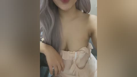 Media: Video of a young woman with long, lavender hair, wearing a strapless, light pink dress with a bow. Her right hand is on her hip, and her left hand is near her chest. The background is blurred, featuring a dark blue wall.