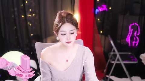 Media: Video of a young East Asian woman with fair skin and brown hair, wearing a light gray off-shoulder sweater, seated in a dimly lit room with black curtains, neon lights, and pink props.