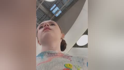 Media: Video of a young woman with light skin, brown hair in a bun, wearing a colorful, abstract-patterned shirt. She gazes upward, surrounded by a modern, industrial-style ceiling with metal grates and circular lights.