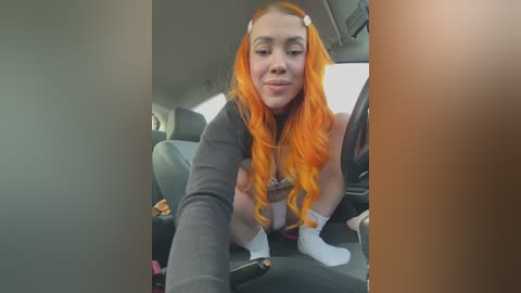 Media: Video of a young woman with vibrant orange hair, wearing a black top, squatting in a car. She has a playful expression, looking out the window.