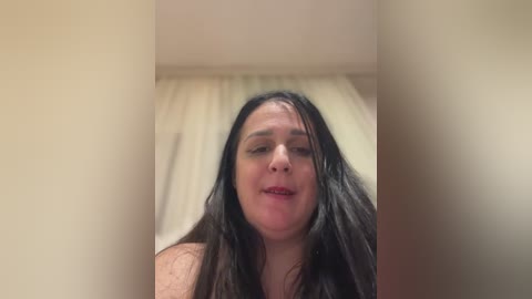 Media: Video of a middle-aged woman with long dark hair, light skin, and a neutral expression. She is indoors, likely in a bedroom, with beige curtains in the background.