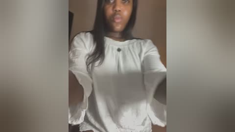 Media: Video of a Black woman with long black hair, wearing a white blouse, standing indoors against a blurred background. She has a pouty expression.