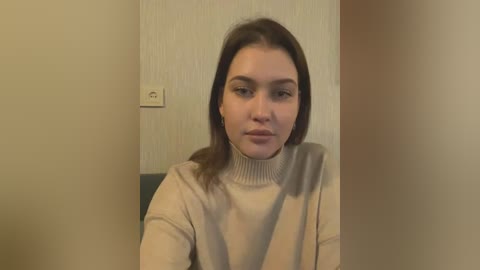 Media: Video of a young woman with fair skin and straight brown hair, wearing a beige turtleneck sweater, seated indoors with beige walls and a light switch in the background.