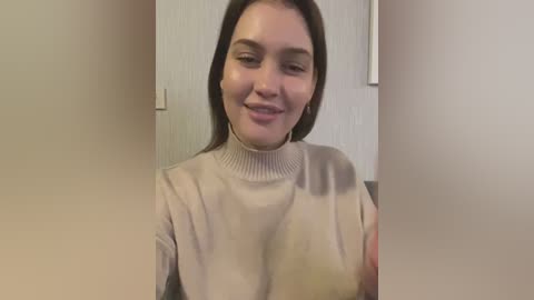 Media: A video of a smiling Caucasian woman with long brown hair, wearing a beige turtleneck sweater, standing in a beige room with a light switch on the wall.