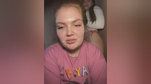 Media: Video of a young woman with pale skin, red hair tied back, wearing a pink \"BYA\" t-shirt, and a playful expression. Another woman is partially visible in the background.