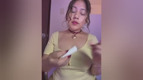 Media: A video of a young woman with medium skin tone, wearing a yellow top and a red choker necklace, holding a white object in a bathroom with purple lighting.