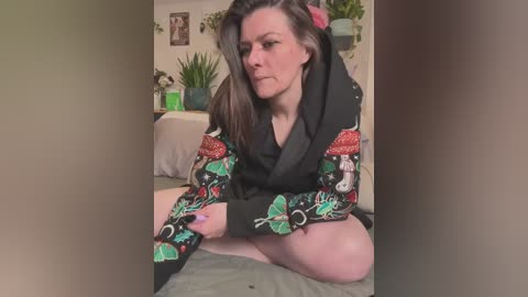 Media: Video of a Caucasian woman with long dark hair, wearing a black robe with colorful Christmas-themed embroidery, sitting cross-legged on a bed in a cozy, dimly lit room with potted plants.