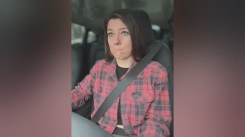 Media: Video of a woman with short brown hair, wearing a red and black plaid shirt, seated in a car with a safety belt across her chest, looking serious.