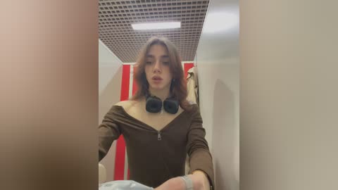 Media: A video of a young woman with shoulder-length brown hair, wearing a brown off-shoulder top, black headphones, and a white bandage on her left wrist, standing in a hallway with white walls and red-striped wallpaper.