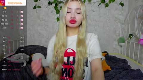 Media: Video of a young, blonde woman with fair skin and red lipstick, wearing a white graphic T-shirt, taking a selfie in a cozy, floral-themed bedroom.