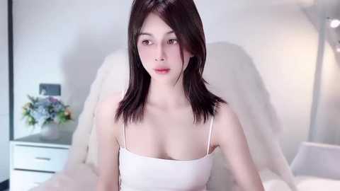 Media: Video of a young Asian woman with straight black hair, fair skin, wearing a white spaghetti-strap top, sitting on a white bed in a minimalist bedroom.