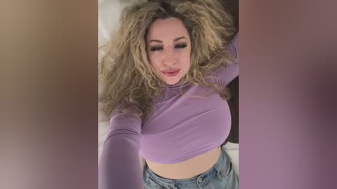 Media: A video of a fair-skinned woman with voluminous, curly blonde hair, wearing a purple long-sleeve crop top and blue jeans, lying on a bed with a soft focus background.