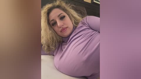 Media: A video of a blonde, plus-sized woman with fair skin, wearing a tight lavender turtleneck, lying on a bed, her large breasts prominently visible. The background is blurred, suggesting an indoor setting.