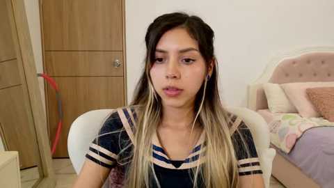 Media: Video of a young Latina woman with long, dark hair and light highlights, wearing a plaid shirt, seated in a white chair in a minimalist bedroom with a bed and wooden door in the background.