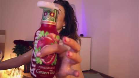 Media: Video of a nude woman holding a tomato-shaped bottle of \"Pomarancio Green Tea\" in a dimly lit room, with a table and flowers visible.