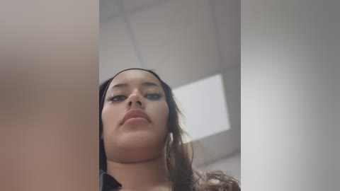 Media: Video of a young woman with light brown skin, long straight brown hair, and full lips, wearing a black top, standing indoors with a white ceiling and a rectangular window.