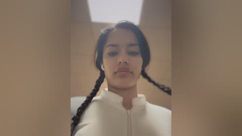 Media: Video of a young woman with medium brown skin and braided black hair, wearing a white, high-collared top, standing in a dimly lit, narrow hallway with beige walls.