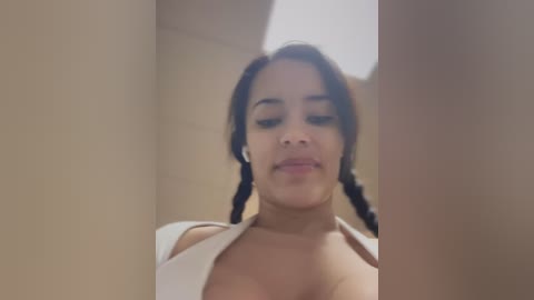 Media: Video of a young woman with medium brown skin, dark braided hair, and a white top. She has a neutral expression, standing in a beige-walled room with a soft, diffused lighting.