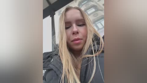 Media: A video of a young blonde woman with fair skin, wearing a dark jacket, looking introspective, with an urban background featuring a metal structure and a building facade.