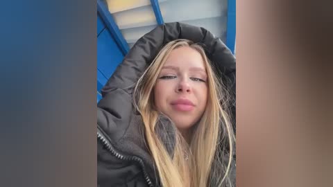 Media: Video of a young Caucasian woman with straight, blonde hair, wearing a black puffy jacket, standing in a blue and white train compartment.