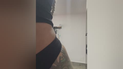 Media: Video of a woman in black underwear, showing a tattooed thigh, in a dimly-lit, white-walled room with a shelf and clothes hanging in the background.