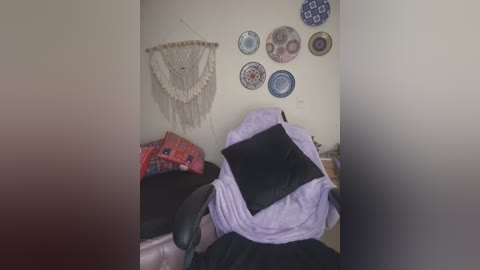 Media: Video of a messy room with a beige wall featuring a macram\u00e9 wall hanging, a purple shawl, and a black pillow on a black chair, surrounded by colorful decorative plates.