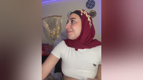 Media: Video of a light-skinned woman with a medium build, wearing a red hijab and white t-shirt, looking to the side in a cozy, dimly-lit room with fairy lights, decorative wall art, and a plush blanket.