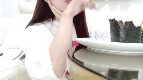 Media: A video of an Asian woman with long, straight brown hair, wearing a white lace dress, holding a fork near a large slice of chocolate cake on a glass pedestal.