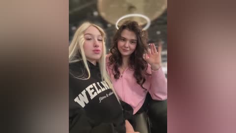 Media: Video of two young women, one with long blonde hair, the other with curly brown hair, wearing black and pink hoodies, respectively. They are seated, with the background blurred.