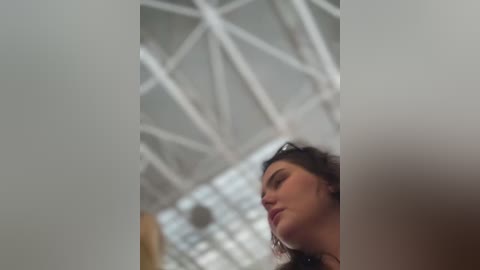 Media: Video of a woman with wavy brown hair, wearing sunglasses, looking upward at a modern, white geometric ceiling with glass panels. She has a neutral expression.