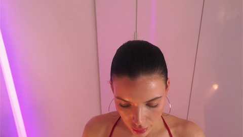 Media: Video of a light-skinned woman with dark hair pulled back, wearing a red halter top, looking down. Background has vertical white panels with pink LED lighting.