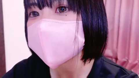 Media: Video of an East Asian woman with straight, black bob haircut, light skin, wearing a pink surgical mask and black clothing, set against a soft pink background.