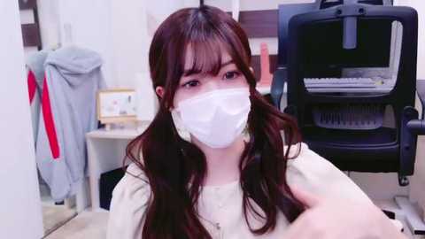 Media: Video of a young Asian woman with long, wavy brown hair, wearing a white surgical mask, light-colored top, and seated in a modern office with a black ergonomic chair and framed certificates on the wall.