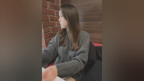 Media: Video of a young woman with straight brown hair, wearing a dark grey knit sweater, seated at a table, looking to the left. Background includes a red brick wall and wooden panels.