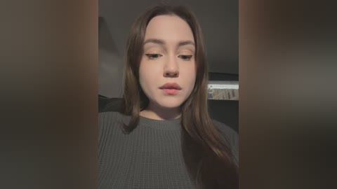 Media: Video of a young woman with long brown hair, wearing a grey ribbed sweater, and looking downward with a neutral expression. The background is dimly lit, featuring a blurred bookshelf.