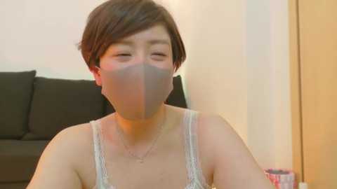 Media: A video of an East Asian woman with short, dark hair and light skin, wearing a white lace camisole and a face mask, sitting on a black sofa in a softly lit room.