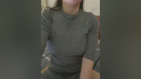 Media: Video of a woman with medium skin tone, wearing a grey long-sleeve top and grey shorts, sitting indoors on a chair with a blurred background.